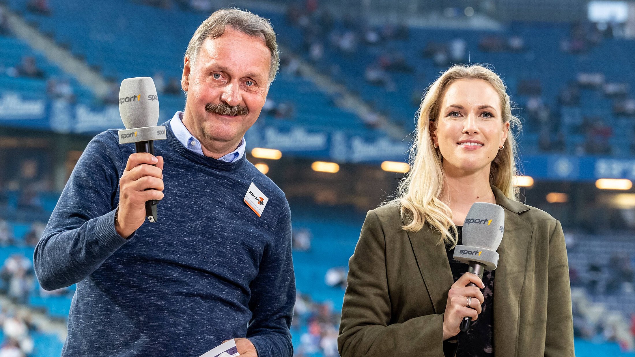 Ruth Hoffmann und Peter Neururer zusammen im Fußballstadion, beide halten ein Mikrofon in der Hand