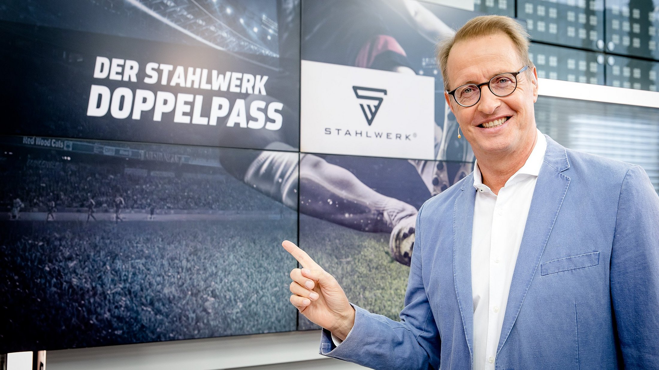 Florian König zeigt auf einen Bildschirm auf dem "Der Stahlwerk Doppelpass" in großer Schrift zu sehen ist