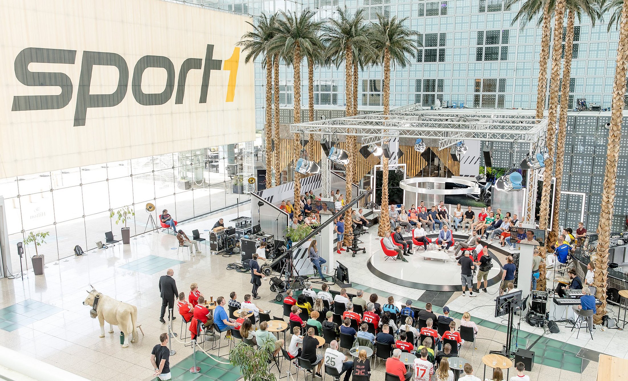 eine Aufnahme von oben. Das Setting vom Doppelpass am Münchner Flughafen, Zuschauer sitzen um ein Podium am Rande sind Palmen und ein sport1 Banner