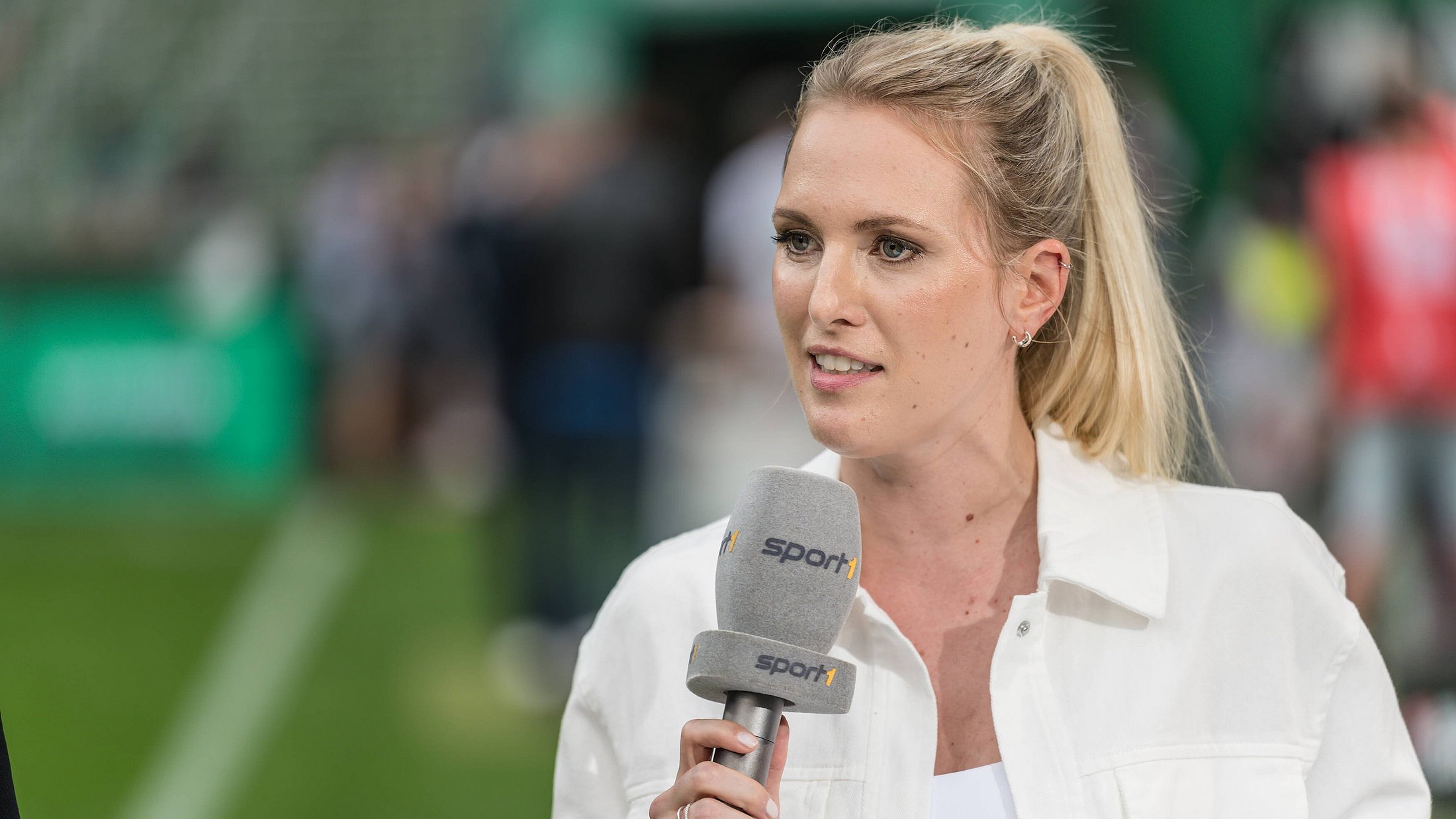 Bild von Ruth Hoffmann (blonde haare, weisse Jacke) mit einem Mikrofon in der Hand in einem Fußballstadion