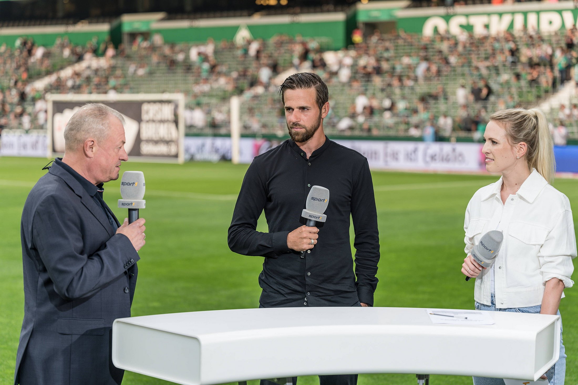 3 Moderatoren stehen an einem Tisch im Fußballstadion